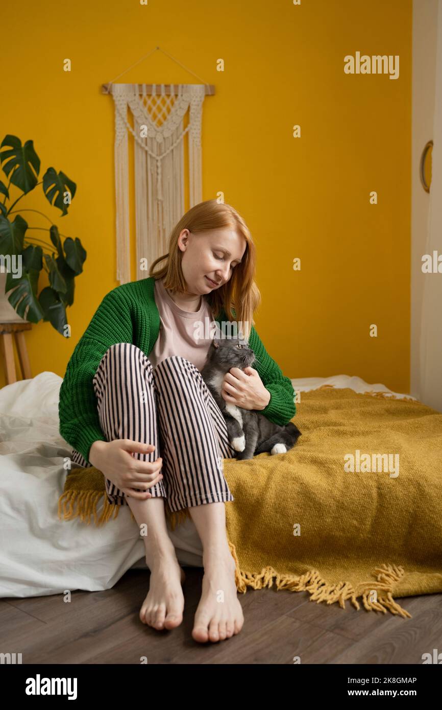 Donna zenzero positivo in pigiama seduta con gli occhi chiusi sul comodo letto carezzando teneramente adorabile gatto con zampe bianche Foto Stock