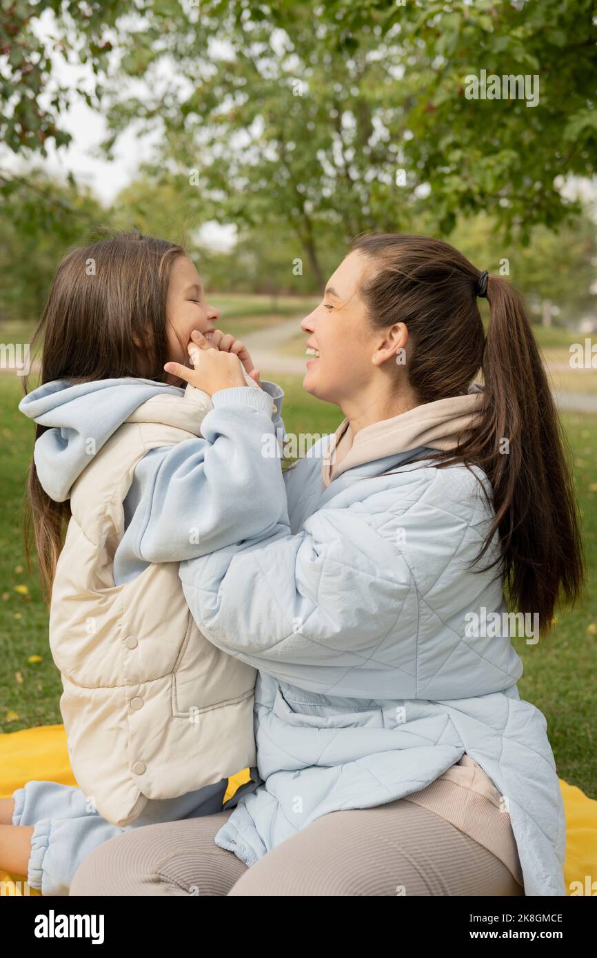 Vista laterale di felice mamma e figlia in outerwear sorridendo e abbracciandosi mentre si siede su coperta gialla il giorno d'autunno nel parco Foto Stock