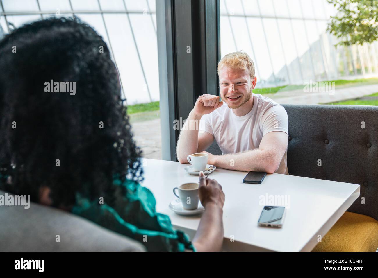 Donna afroamericana tagliata irriconoscibile con allegro bionda uomo in t shirt bianca seduta a tavolo con tazze di caffè e guardando ciascuno Foto Stock