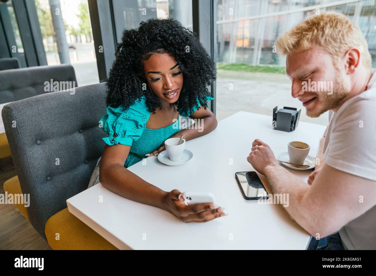 Un uomo delizioso e una ragazza nera positiva seduti insieme al tavolo della caffetteria che naviga sullo smartphone Foto Stock