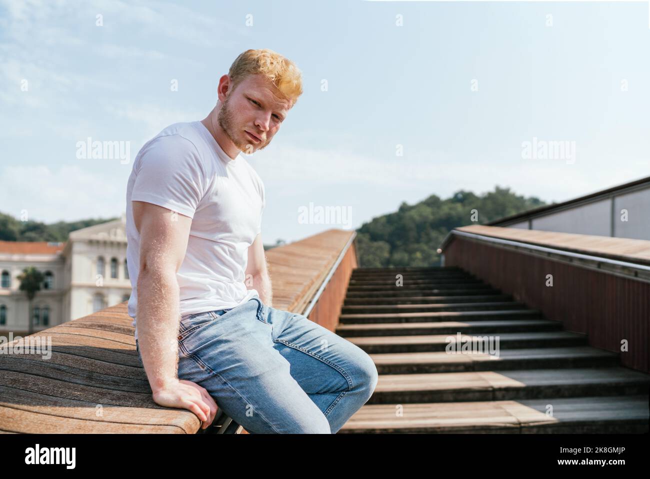 Vista laterale del giovane uomo biondo muscoloso bearded in t shirt bianca e jeans seduti sul corrimano di scale di legno contro la città sfocata e guardando venuto Foto Stock