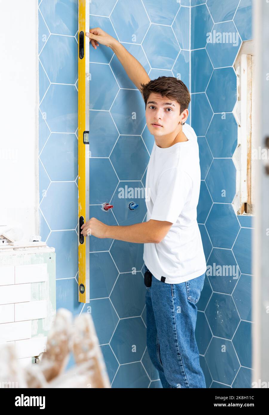 Giovane uomo che controlla la qualità delle piastrelle posate a parete con misuratore di livello Foto Stock