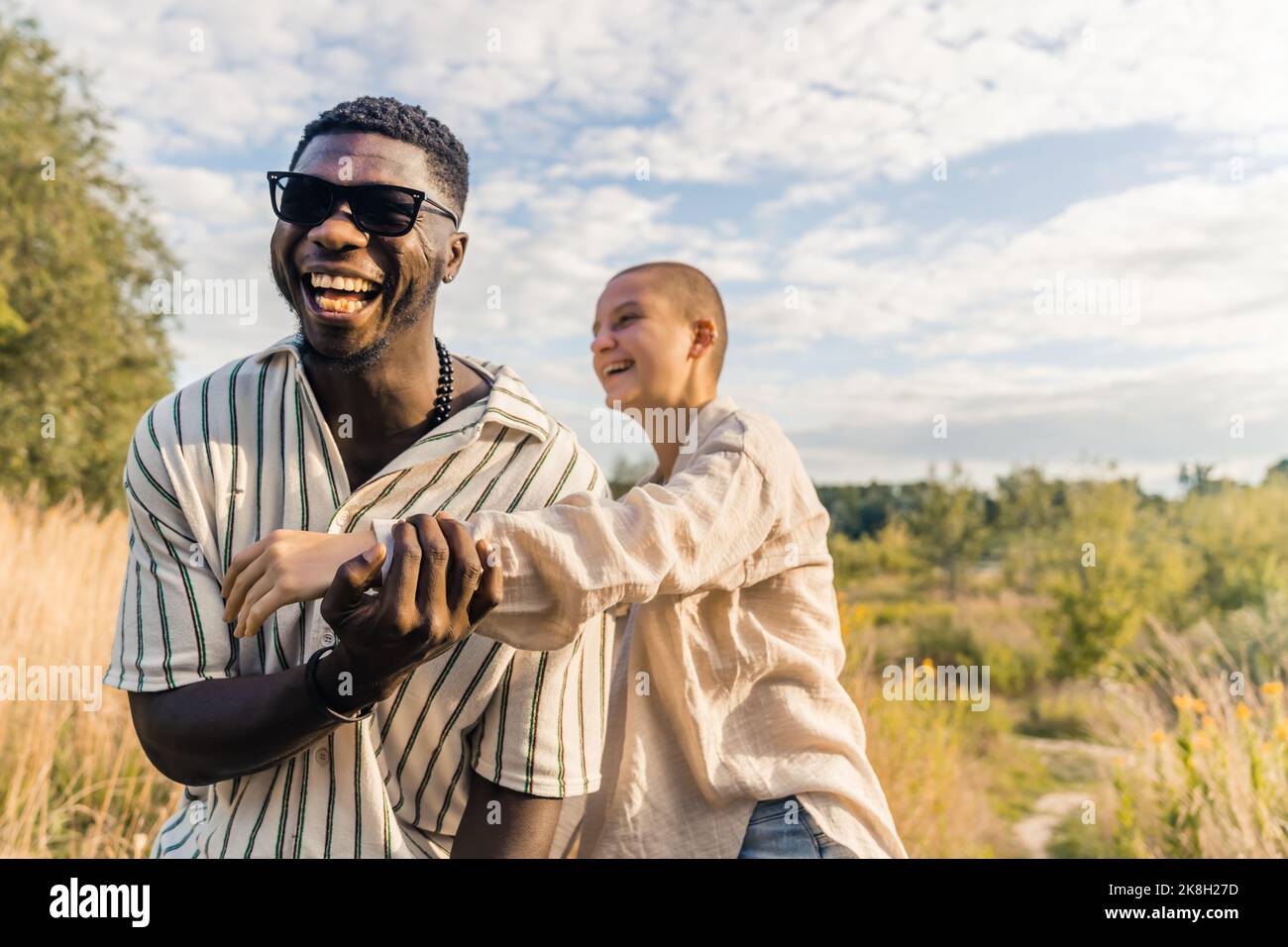 Divertimento e risate con gli amici e quelli vicini. Coppia multietnica che si diverte in campagna. Ridendo uomo nero in abbigliamento casual toccando il braccio del suo calvo amico caucasico. Foto di alta qualità Foto Stock