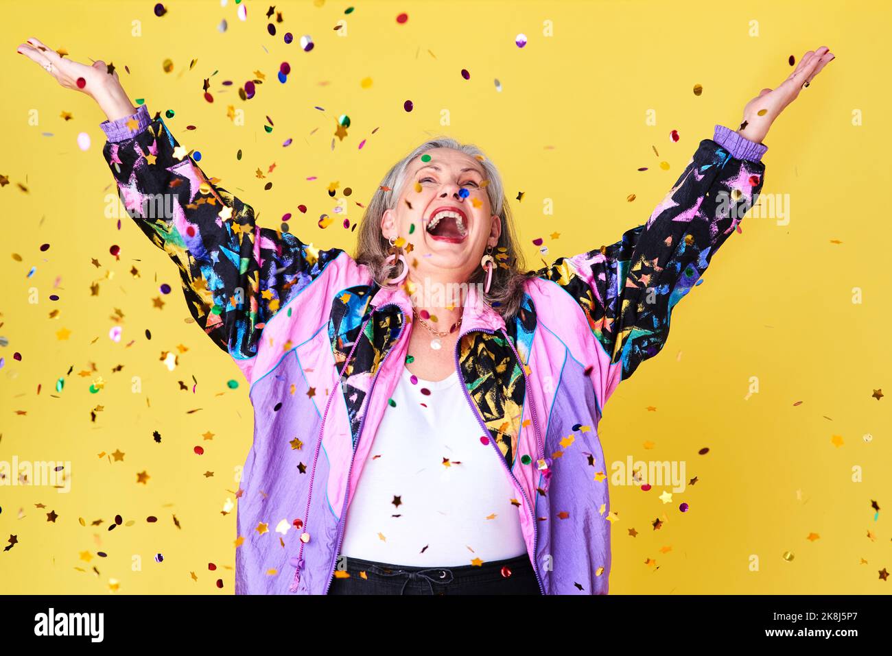 La pioggia di confetti è il mio clima preferito: Una donna anziana allegra ed elegante che festeggia con i confetti che cadono sopra di lei contro un giallo Foto Stock
