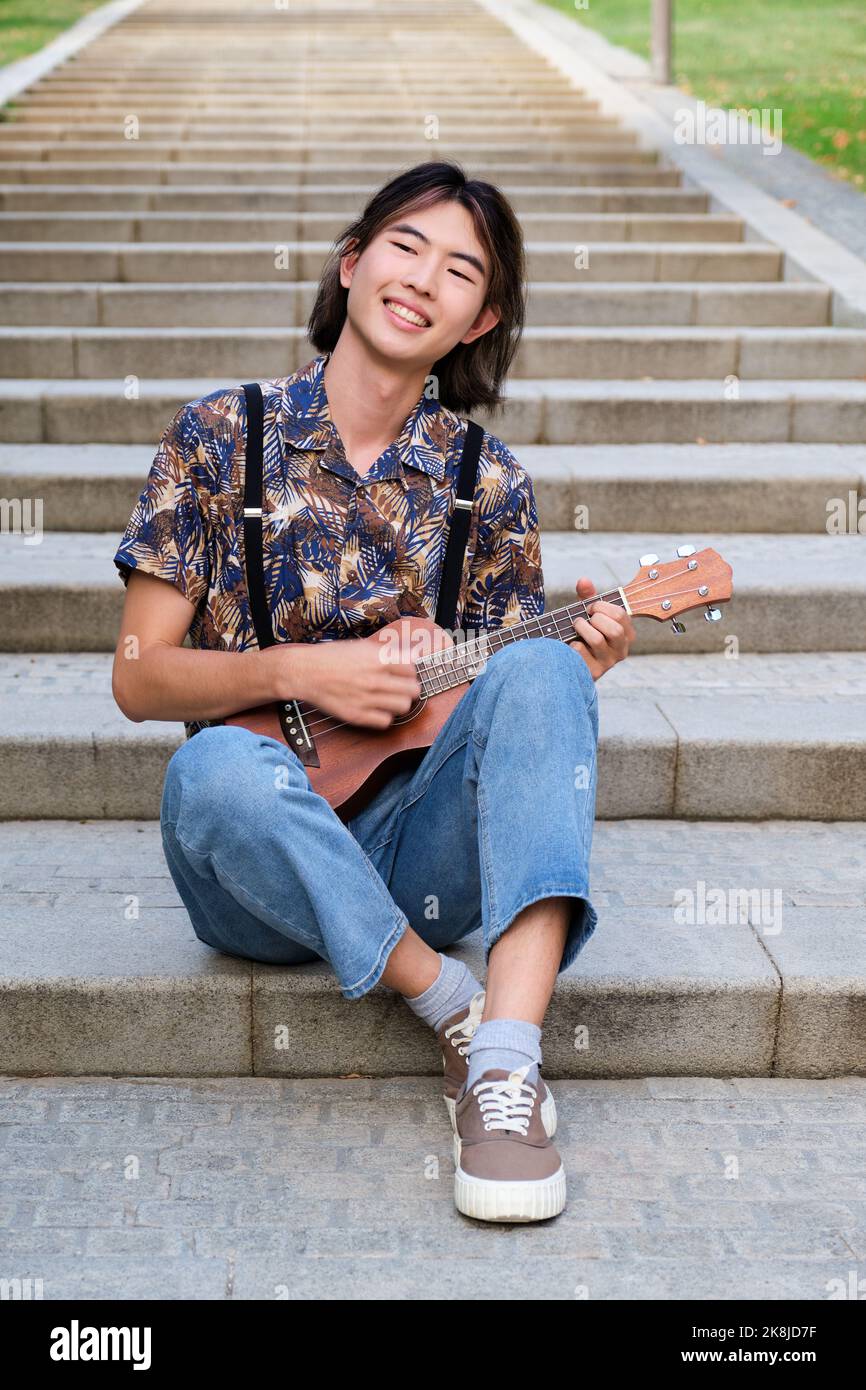 Ragazzo asiatico sorridente e suona la chitarra acustica Ukulele seduto sulle scale. Foto Stock