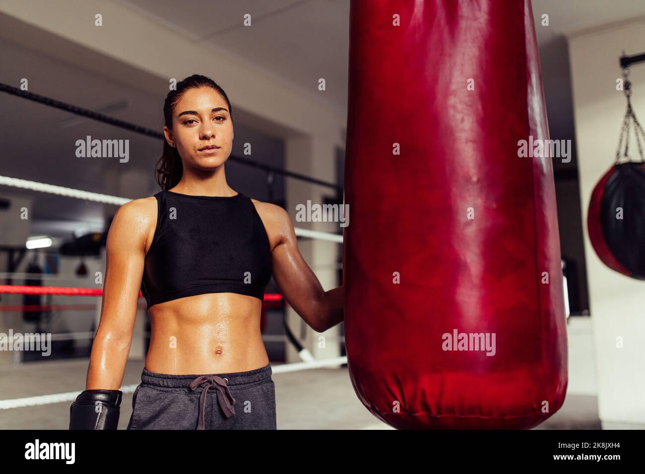 Pugile femminile che guarda la macchina fotografica mentre si trova accanto a una borsa da punzonatura in una palestra fitness. Giovane donna atletica che indossa abbigliamento sportivo e glo di boxe Foto Stock