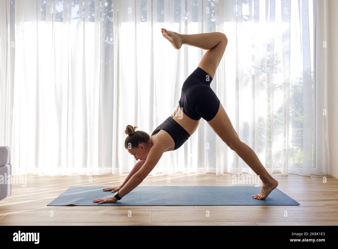 Giovane donna in abbigliamento sportivo nero che fa yoga in camera con grande finestra Foto Stock
