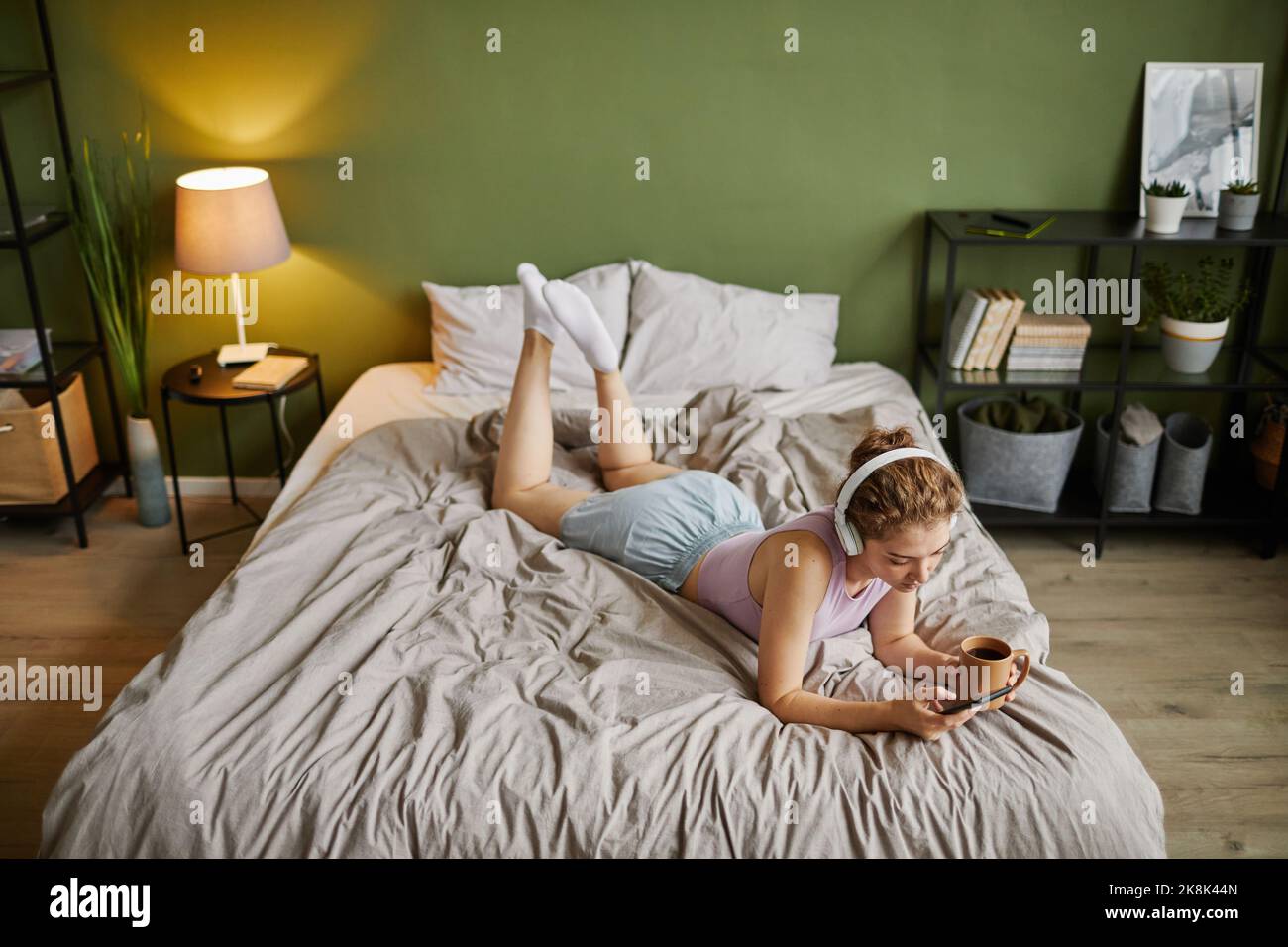 Vista ad alto angolo della giovane donna con le cuffie utilizzando il telefono cellulare mentre sdraiata sul letto con una tazza di caffè Foto Stock