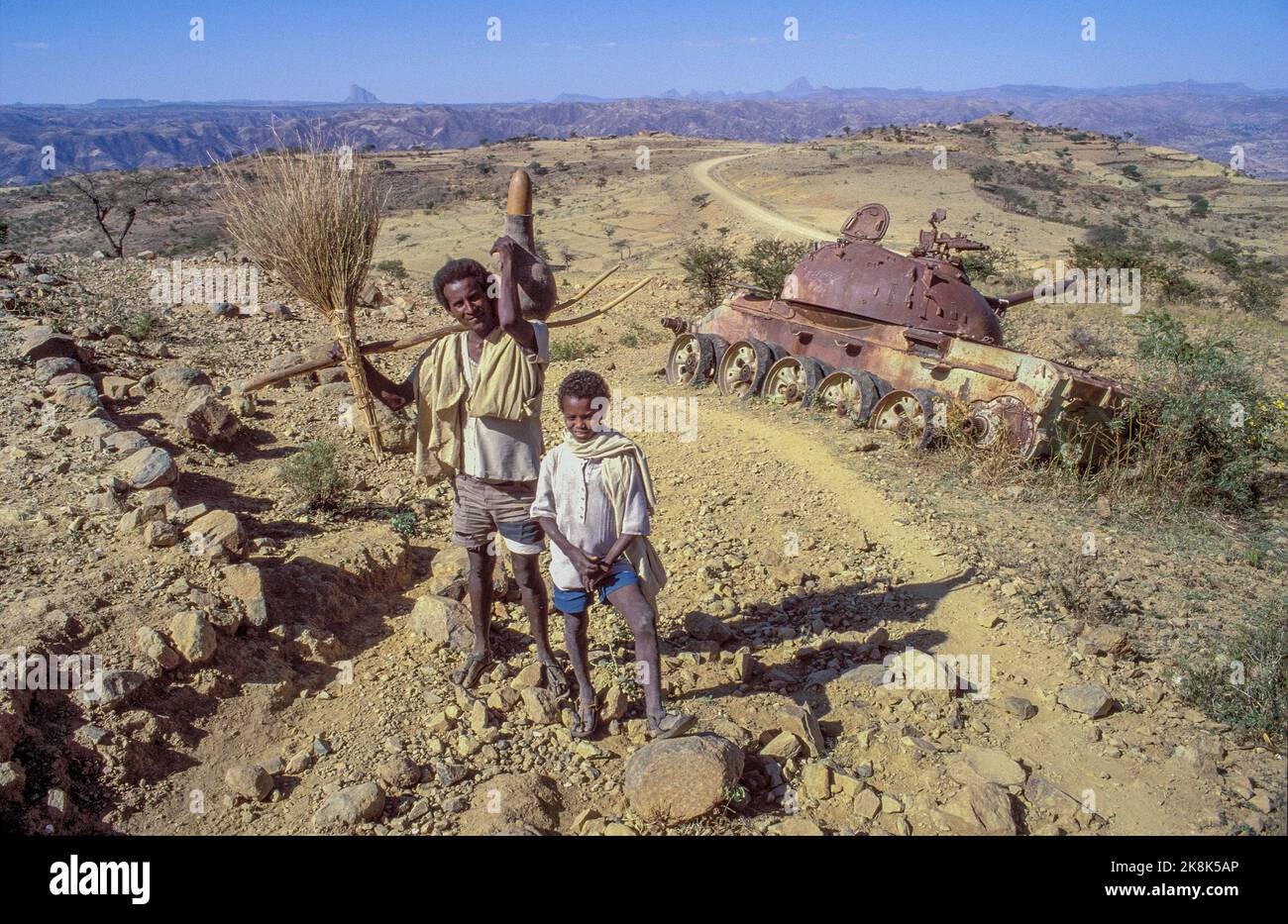 Etiopia, Tigray; padre e sole hanno appena lasciato il loro lavoro agricolo sul campo e passano un veicolo blindato abbandonato e sdraiato nel deserto. Un rem Foto Stock