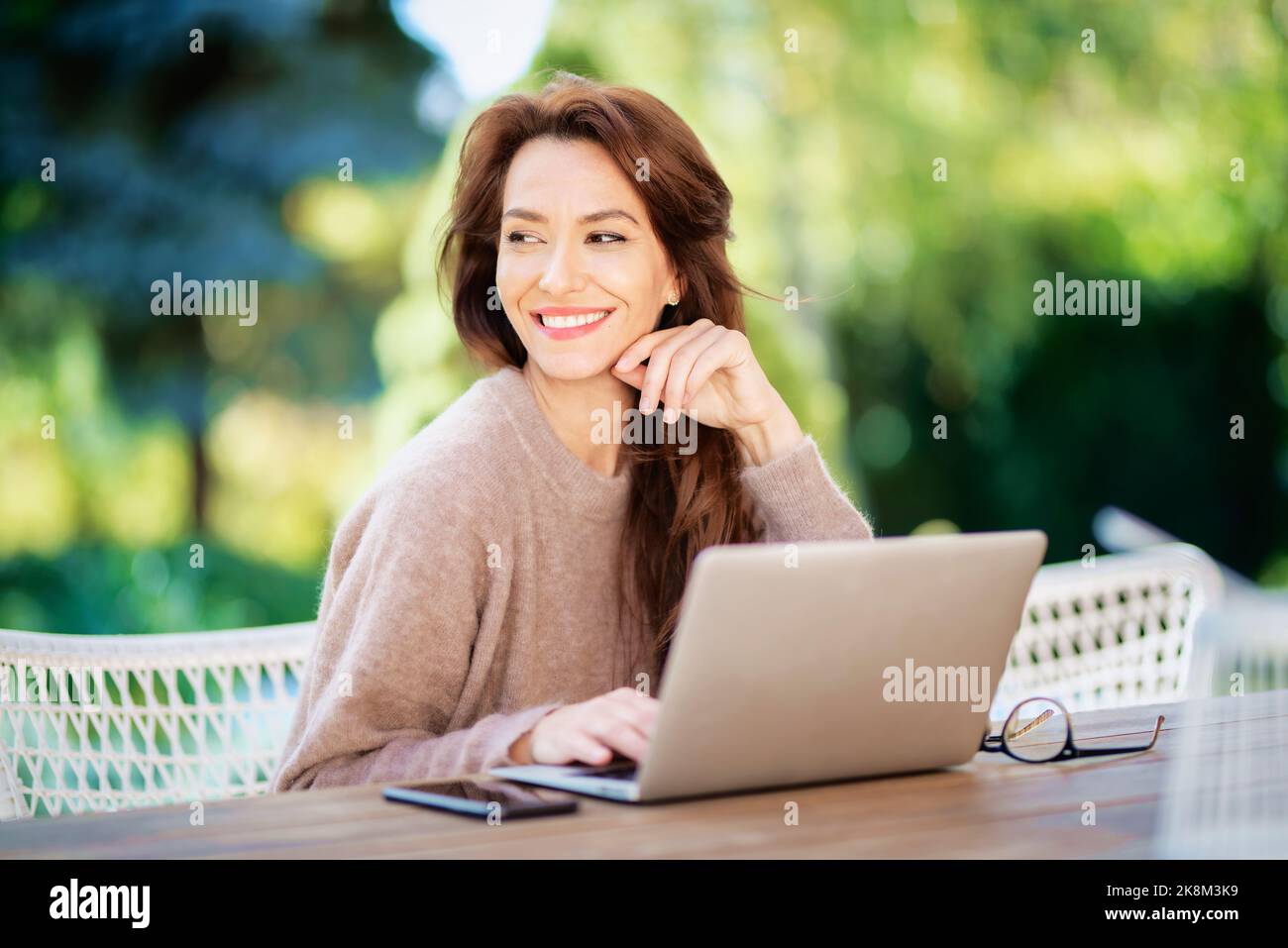 Donna di mezza età con notebook seduto sul balcone. Attraente donna che indossa maglione e sorridente allegro. Foto Stock