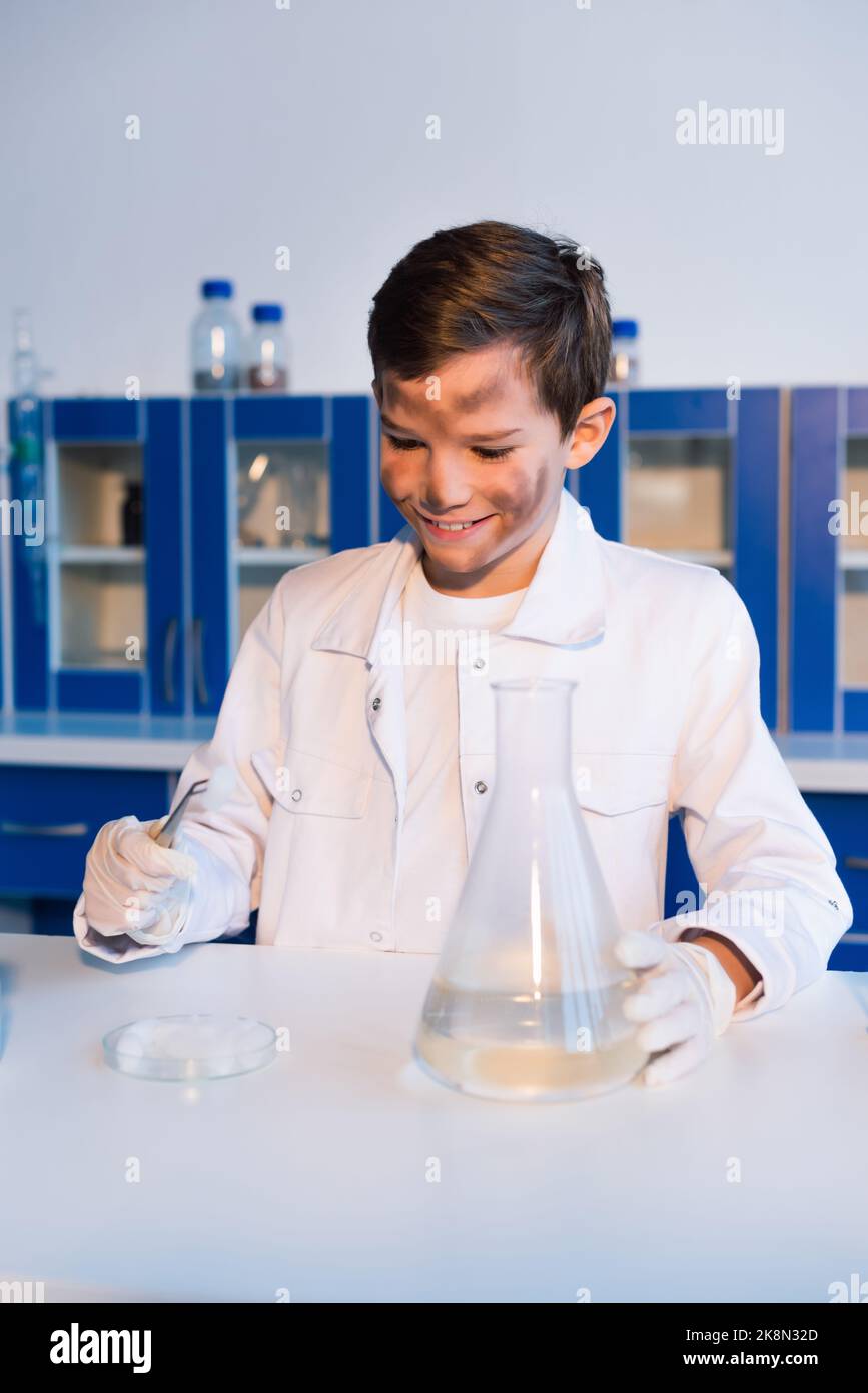 ragazzo sorridente con il viso sporco che tiene le pinzette vicino alla capsula di petri e alla fiasca in laboratorio, immagine stock Foto Stock