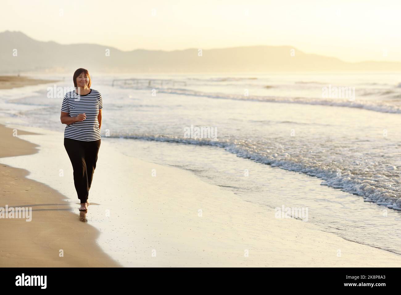 Donna sorridente di mezza età che corre sulla spiaggia all'alba. 40s o 50s bella signora matura in abbigliamento sportivo facendo jogging allenamento godendo di fitness Foto Stock