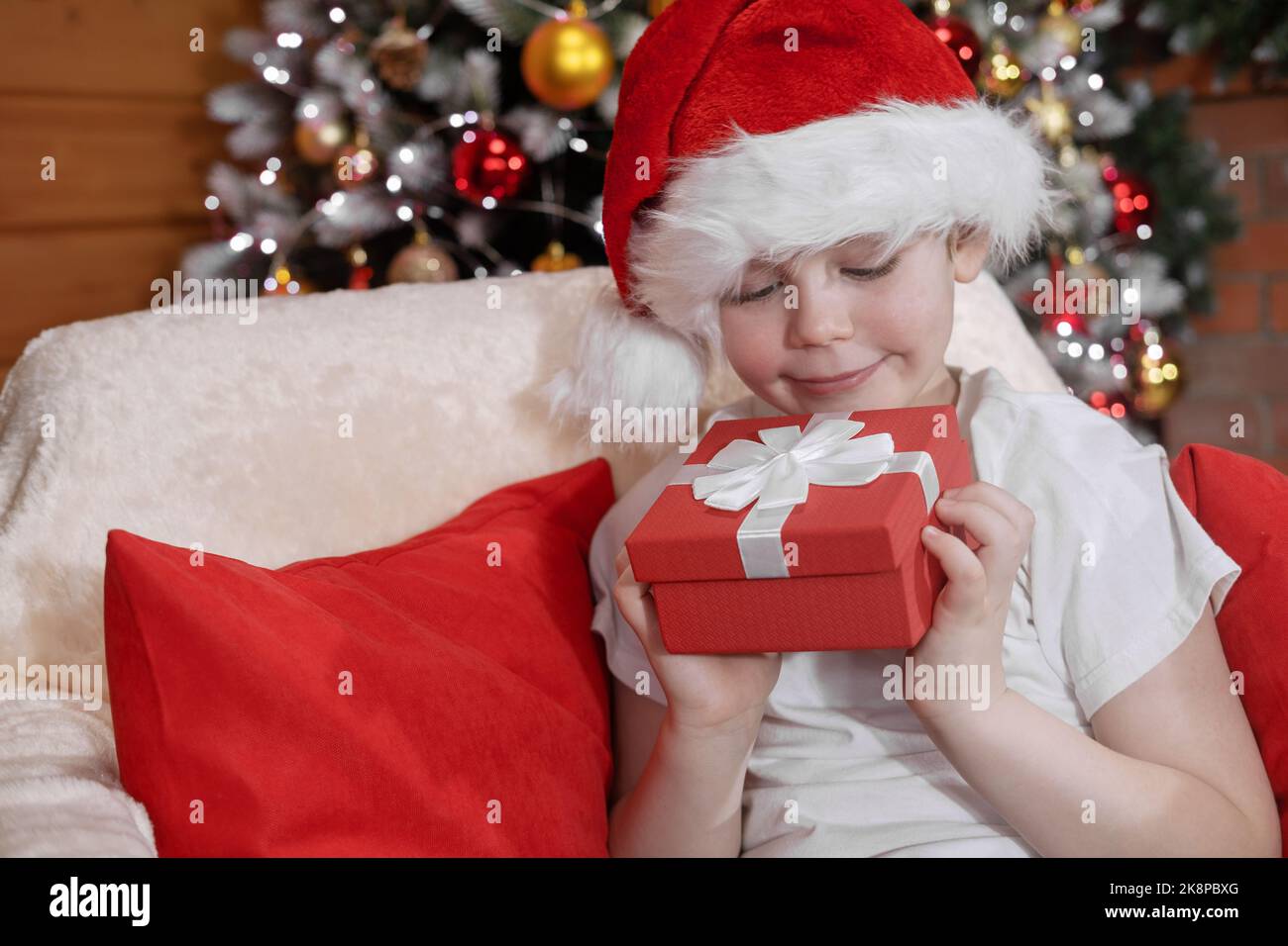 Buon Natale bambini. Un ragazzo in un cappello di Babbo Natale con un regalo di Babbo Natale, vicino a un albero di Natale con luci di Natale e un camino. Natale in famiglia Foto Stock