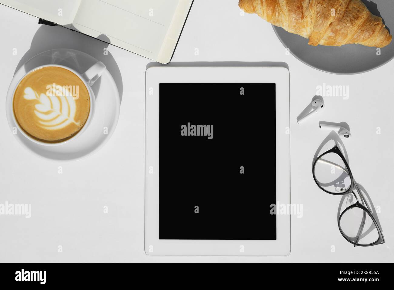 Vista dall'alto di caffè, croissant, tavoletta, blocco note e occhiali sullo sfondo bianco della scrivania. Foto Stock
