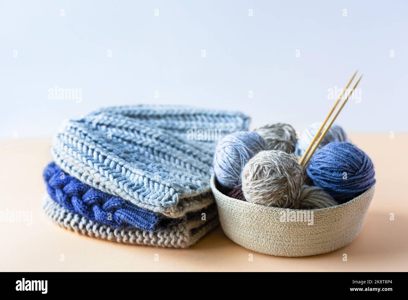 Palle grigie, marroni e blu di fili da maglieria in un cesto beige con aghi da maglieria in bambù e una pila di cappelli a maglia per inverno e autunno, maglia Foto Stock