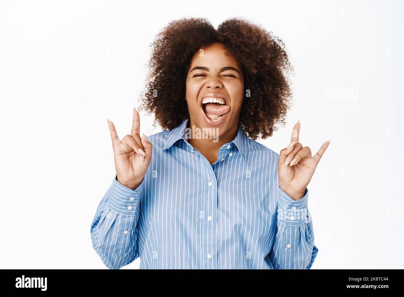Donna afro-americana spensierata, mostra rock on, pesante gesto di metallo e sorridente, esprimere positività e felicità, in piedi su sfondo bianco Foto Stock