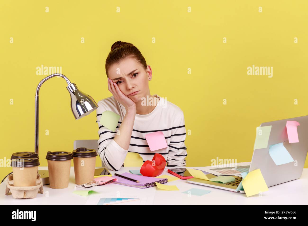 Annoiato donna impiegato ufficio coperto con appiccicoso seduta sul posto di lavoro appoggiando testa sulle mani, solo deluso donna stanca di lavoro wimmp. Note Studio interno girato isolato su sfondo giallo Foto Stock
