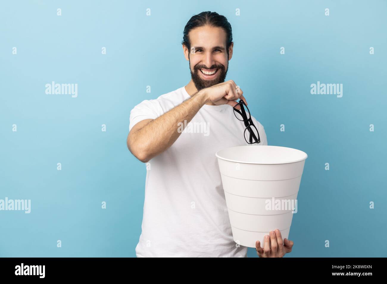 Ritratto dell'uomo con la barba che indossa una T-shirt bianca che getta gli occhiali nel bidone della spazzatura, tratta la sua vista, guardando sorridendo alla fotocamera. Studio in interni isolato su sfondo blu. Foto Stock