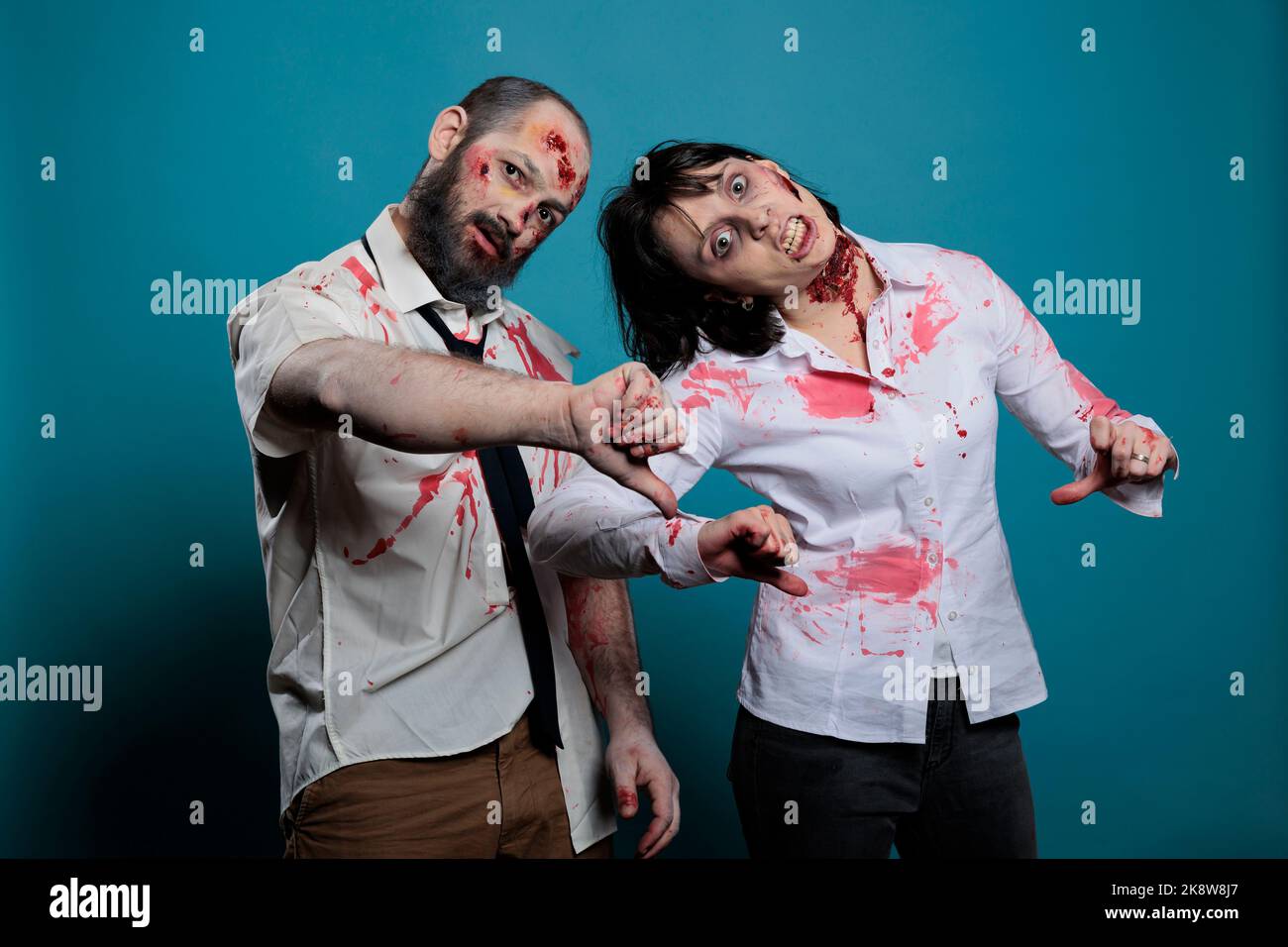Mostri di Halloween che mostrano i pollici giù sulla macchina fotografica, esprimendo disapprovazione e dispiacere in studio. Coppia di cervello che mangia zombie con cicatrici sanguinose e ferite brutte, diavoli spaventosi. Foto Stock