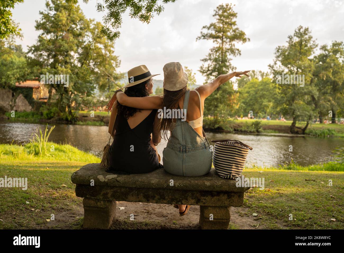 due amiche con cappelli che chiacchierano seduti su una panca di pietra di fronte al fiume Foto Stock