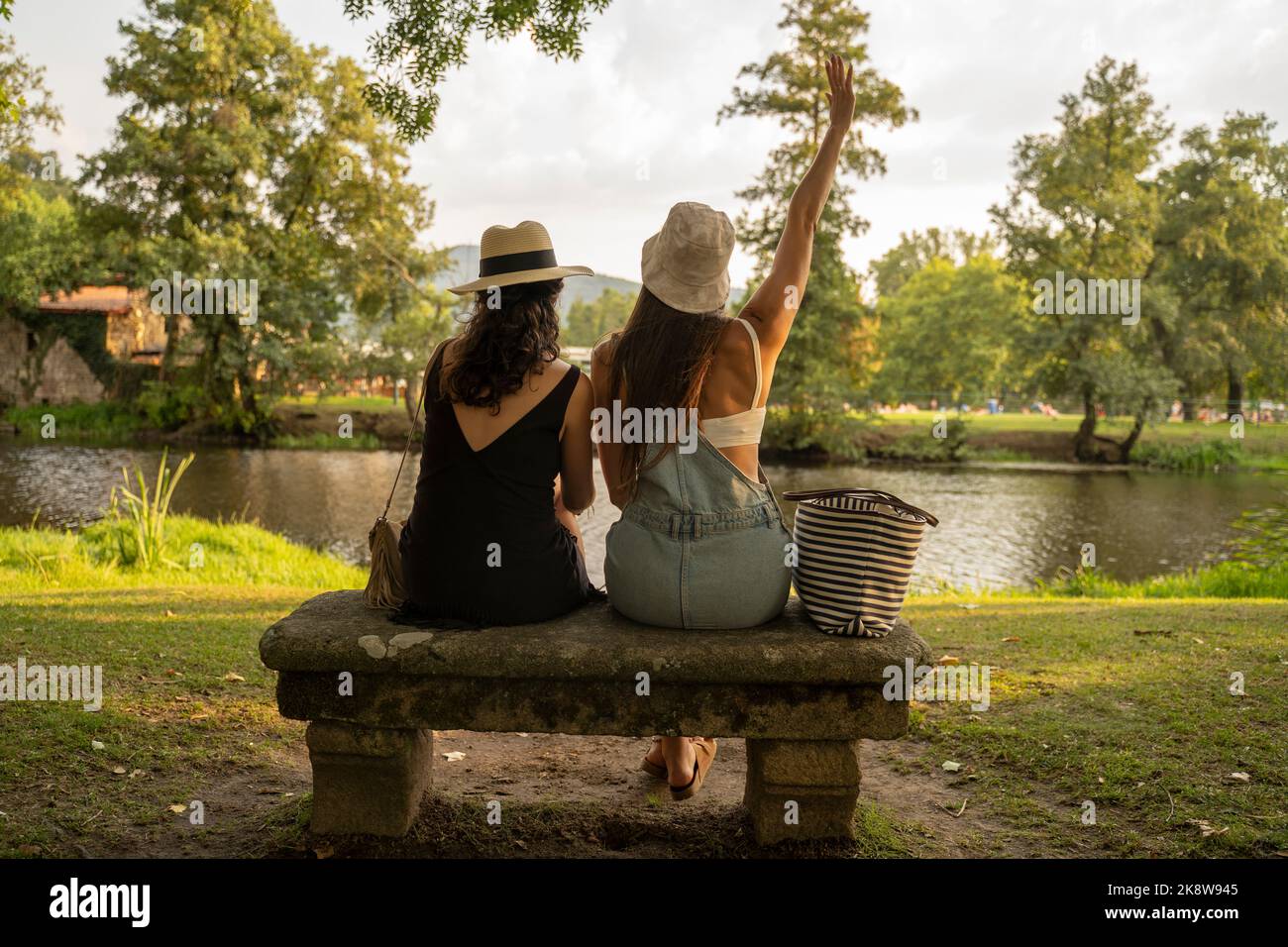 due amiche con cappelli che chiacchierano seduti su una panca di pietra di fronte al fiume Foto Stock