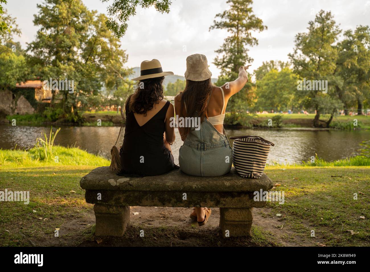 due amiche con cappelli che chiacchierano seduti su una panca di pietra di fronte al fiume Foto Stock