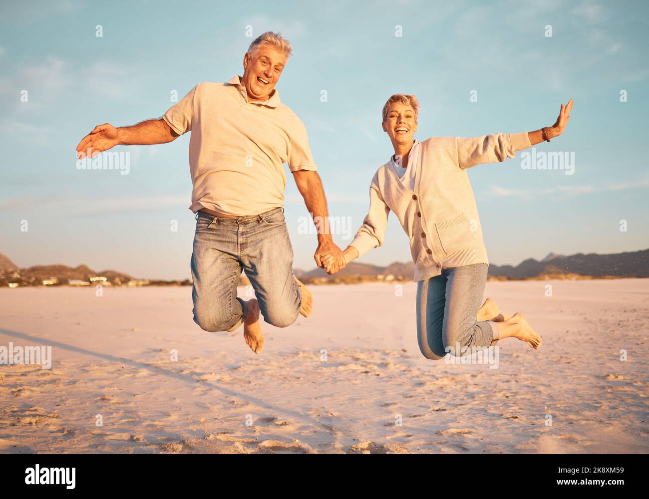 La coppia, la spiaggia e gli anziani attivi tengono le mani mentre saltano nella sabbia, felici ed eccitati al tramonto. Amore, famiglia e libertà con uomo e donna maturi Foto Stock