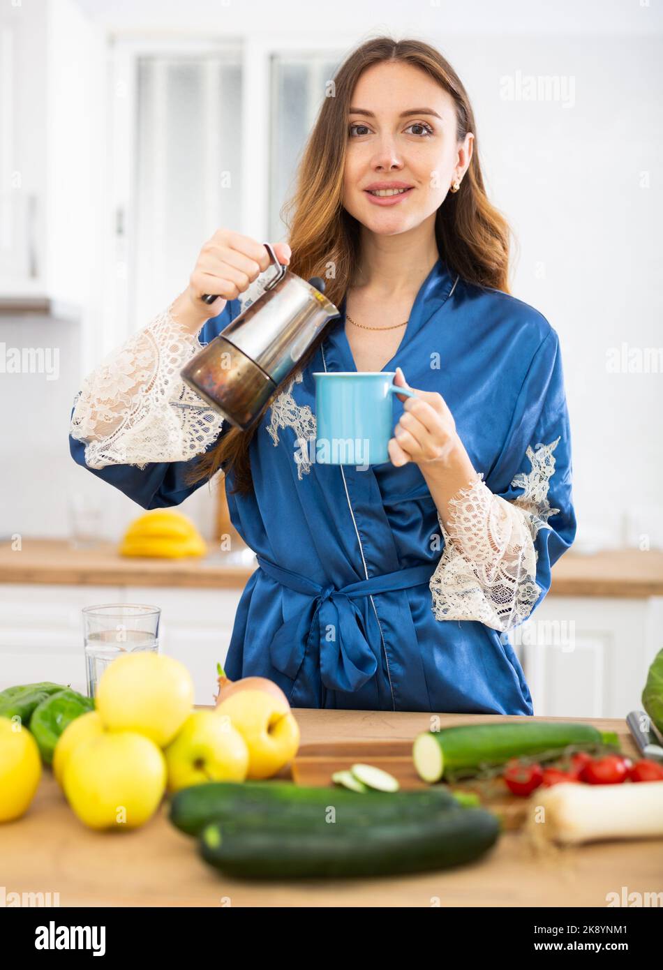 Giovane donna sorridente in vestito blu che versa caffè in tazza in cucina a casa Foto Stock