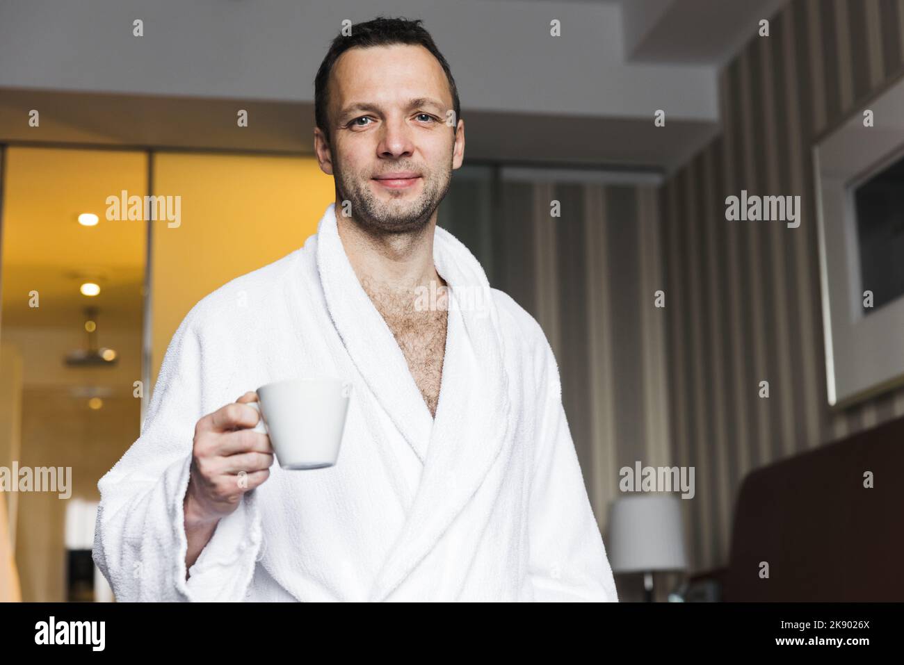 Giovane uomo caucasico sorridente in un accappatoio di cotone bianco in piedi con una tazza di caffè in una camera d'albergo Foto Stock