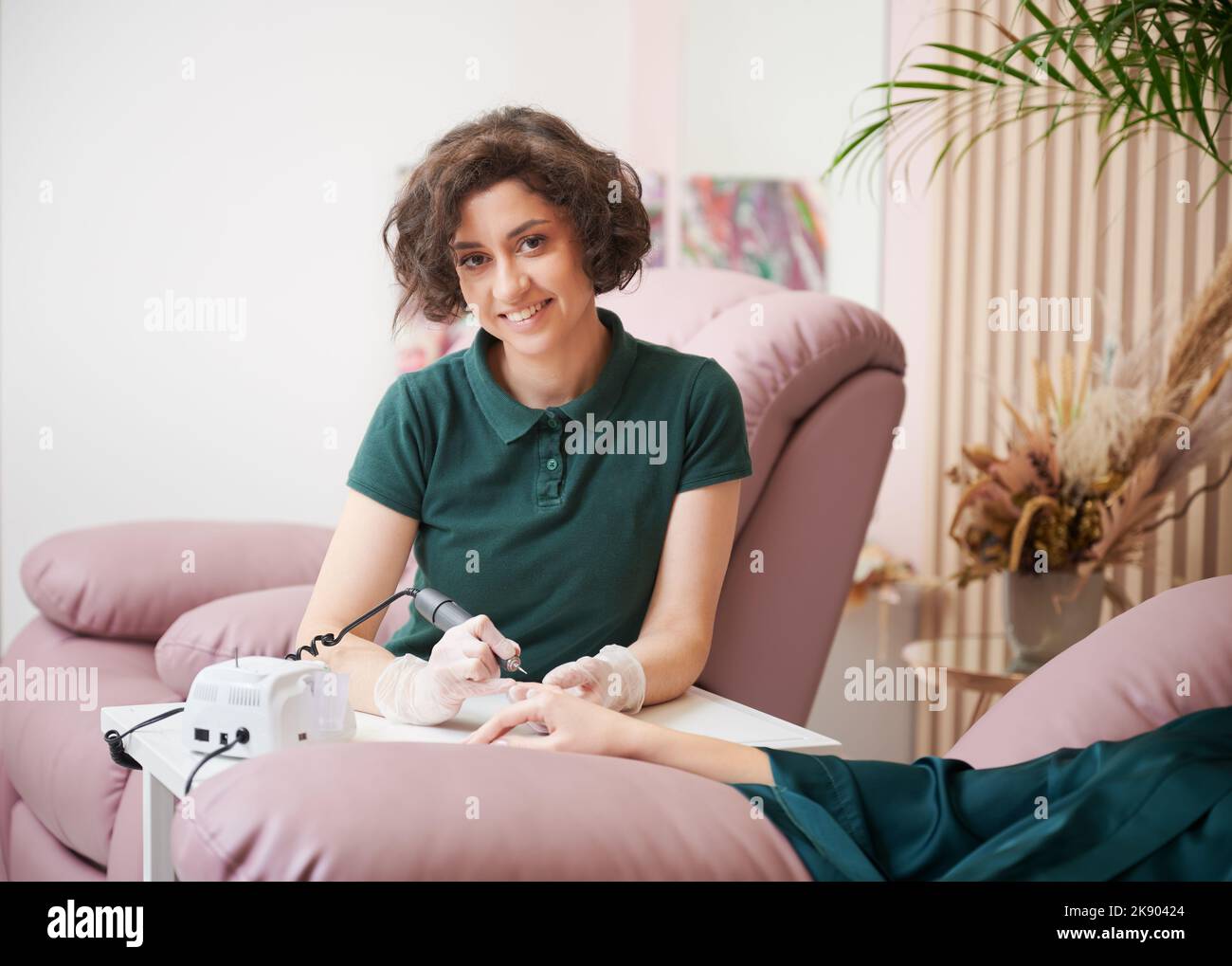 Ritratto del maestro di manicure che tiene la mano del cliente e che utilizza un trapano elettrico per unghie. Donna felice manicure guardando la macchina fotografica e sorridente mentre fa manicure per la donna in salone di bellezza. Foto Stock