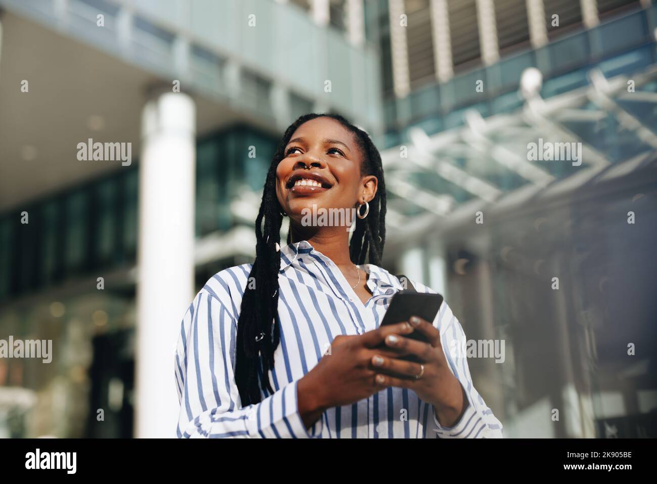 Giovane e premurosa donna d'affari che guarda via con un sorriso mentre si è in viaggio per lavorare in città. Felice giovane donna d'affari che usa uno smartphone durante il suo viaggio Foto Stock