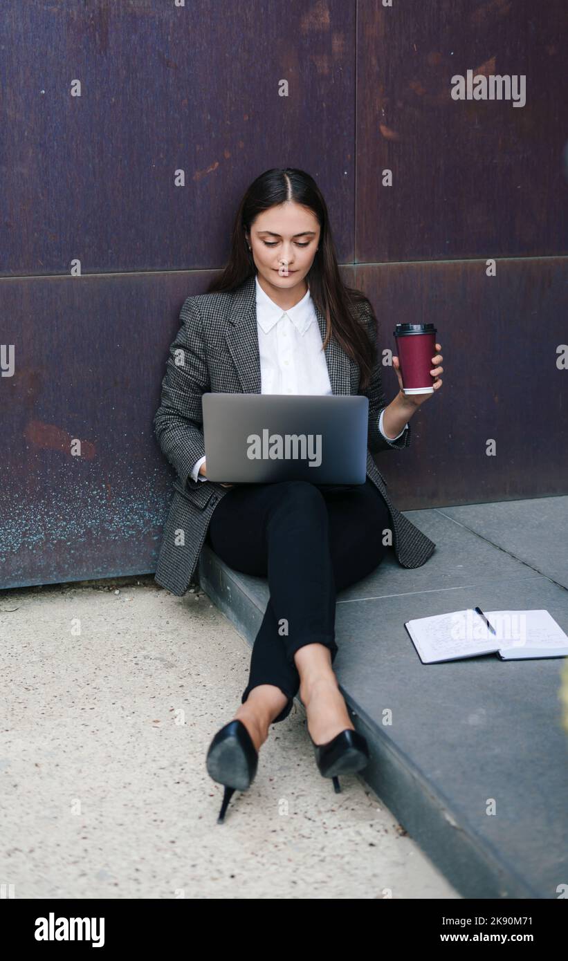 Vista frontale ritratto di una bella donna di successo seduta all'aperto ufficio, utilizzando computer portatile e bere caffè alla luce del mattino. Stile di vita e. Foto Stock
