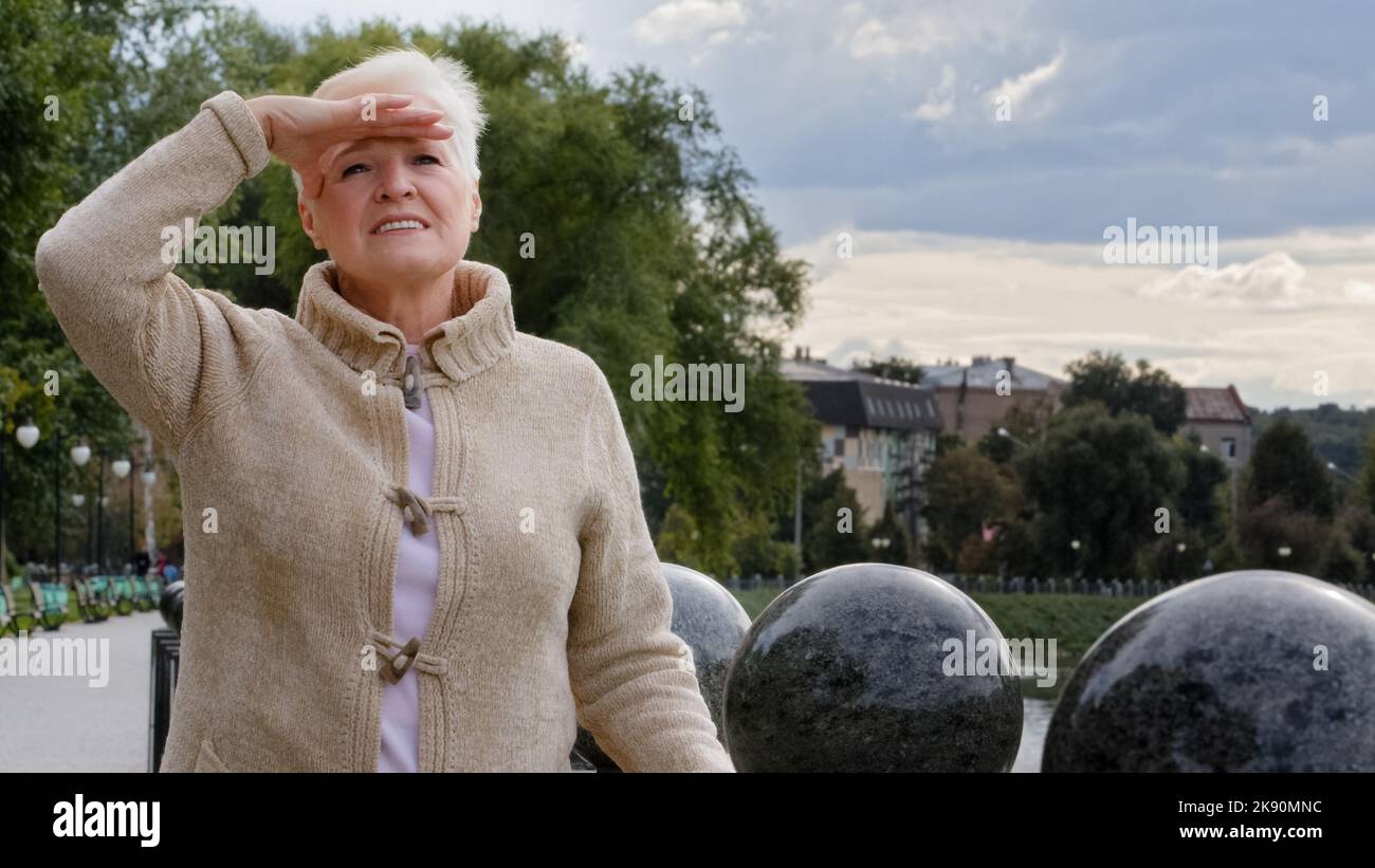 Attraente donna anziana mette mano a fronte, coprendo gli occhi dalla luce solare brillante, peering in distanza, cercando di vedere qualcuno. Elegante pensione Foto Stock