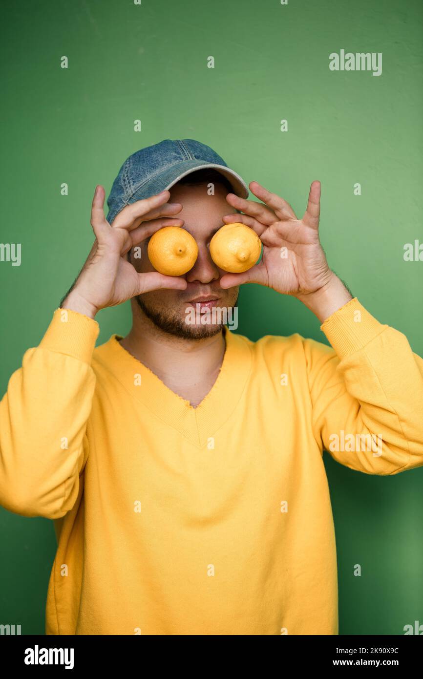 Giovane con la barba in un cappello sulla testa con i limoni sugli occhi su uno sfondo verde. Ritratto di un ragazzo in un maglione giallo Foto Stock