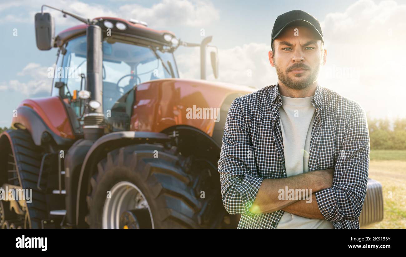 Un contadino bearded in un berretto e una maglietta a quadri si trova davanti a un trattore agricolo Foto Stock
