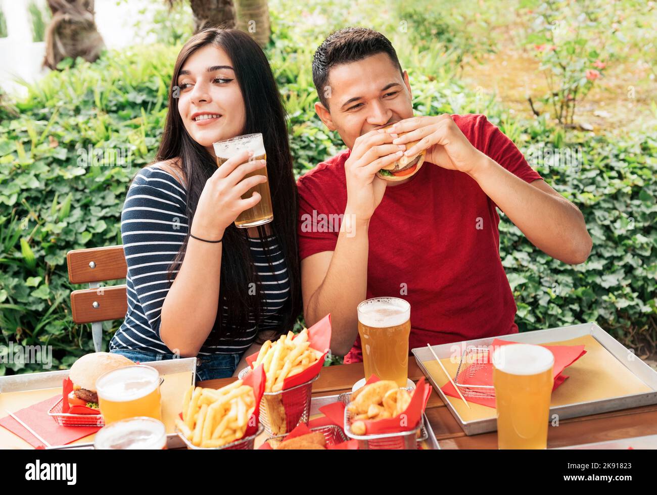 Coppia mangiando gustosi hamburger e bevendo birra alcolica mentre si siede sulla terrazza del caffè all'aperto con piante verdi in città Foto Stock