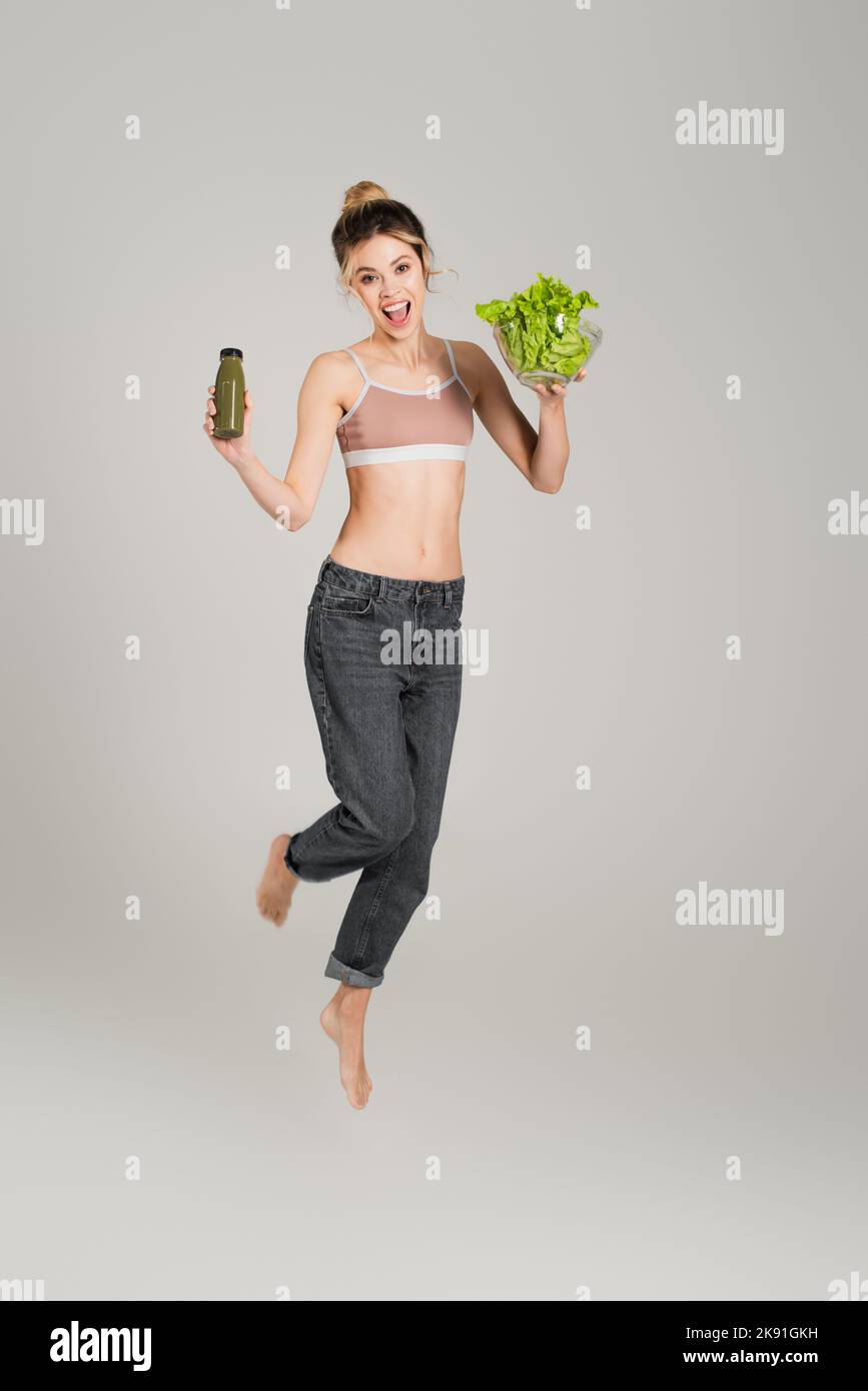 donna emozionata a piedi nudi con corpo sottile che si livella con lattuga e bottiglia di frullato su sfondo grigio Foto Stock