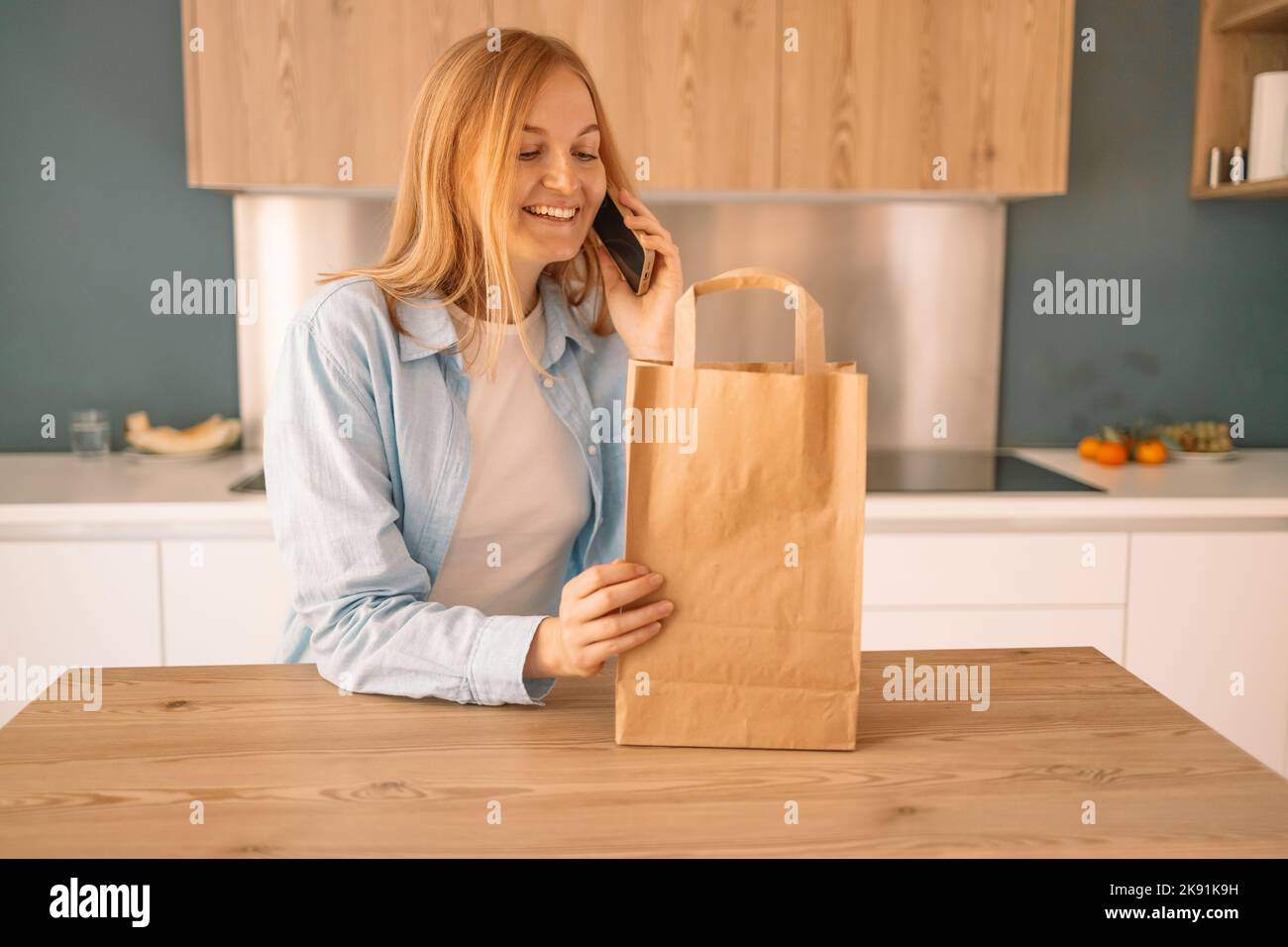 Bella giovane donna caucasica tenere eco shopping borsa dopo lo shopping con smartphone in cucina moderna a casa Foto Stock