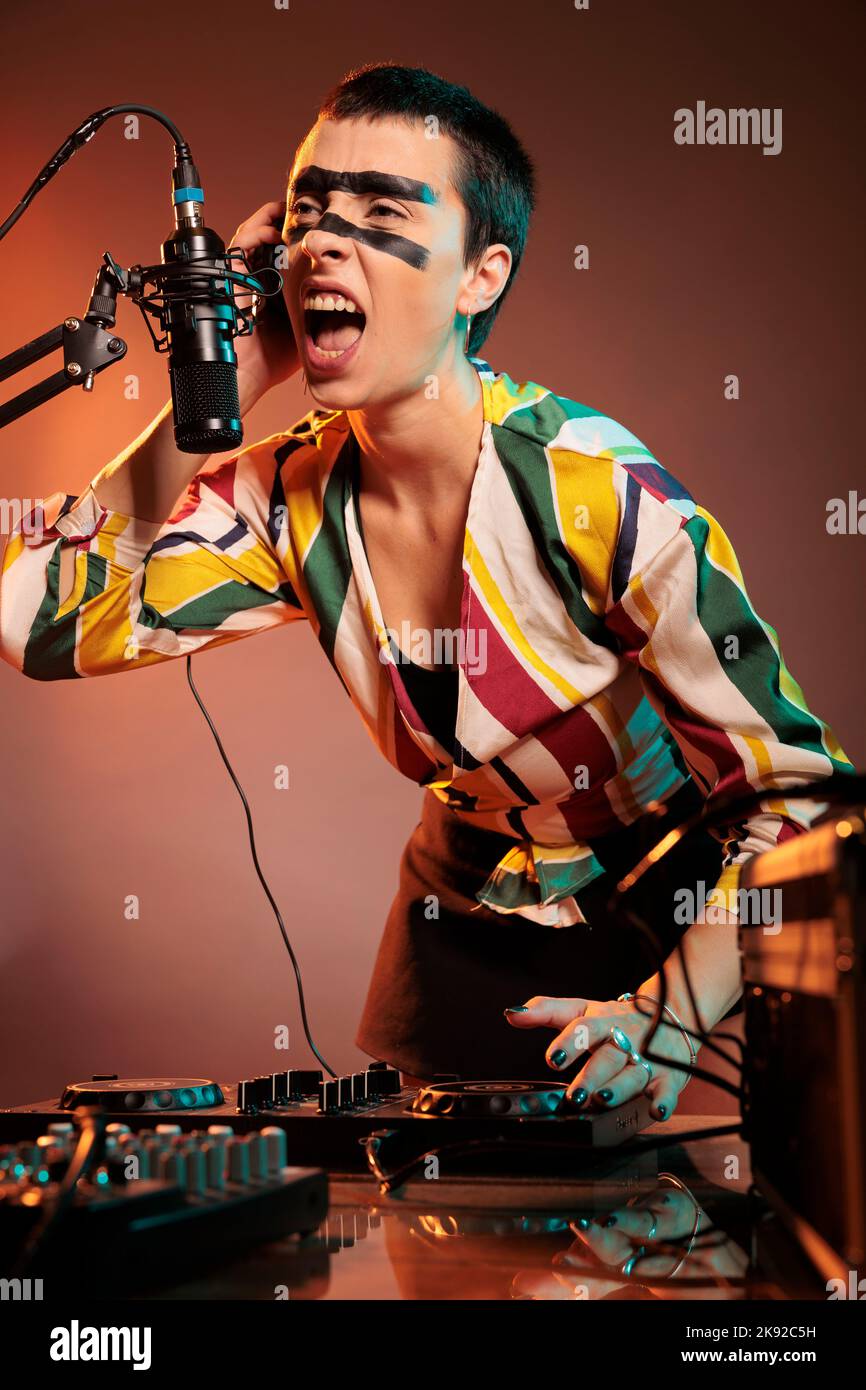 Musicista che urla al microfono, mixando musica techno ai giradischi dj su sfondo fumoso. Artista femminile che suona suoni a strumenti audio e mixer stereo. Foto Stock