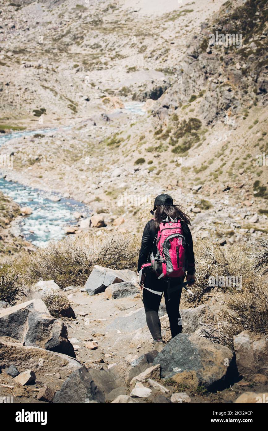 Vista posteriore di una donna escursionista che cammina attraverso un sentiero roccioso accanto a un fiume di montagna, con uno zaino sopra. Scatto verticale Foto Stock