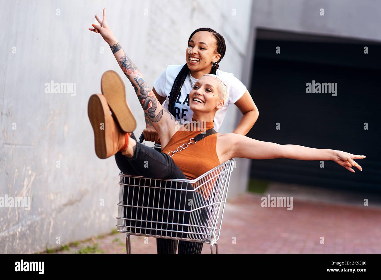 Im avere buon divertimento. Una giovane donna energica che spinge la sua amica femmina in un carrello di shopping all'aperto. Foto Stock