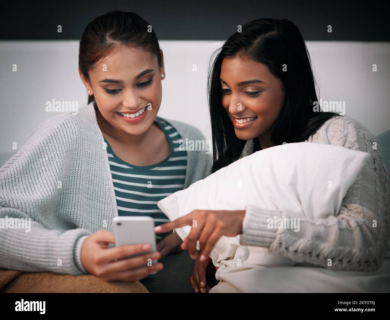Scarica questa app per l'editing di immagini, sembra divertente. Una giovane donna che mostra qualcosa alla sua amica sul cellulare mentre si siede insieme a un letto. Foto Stock