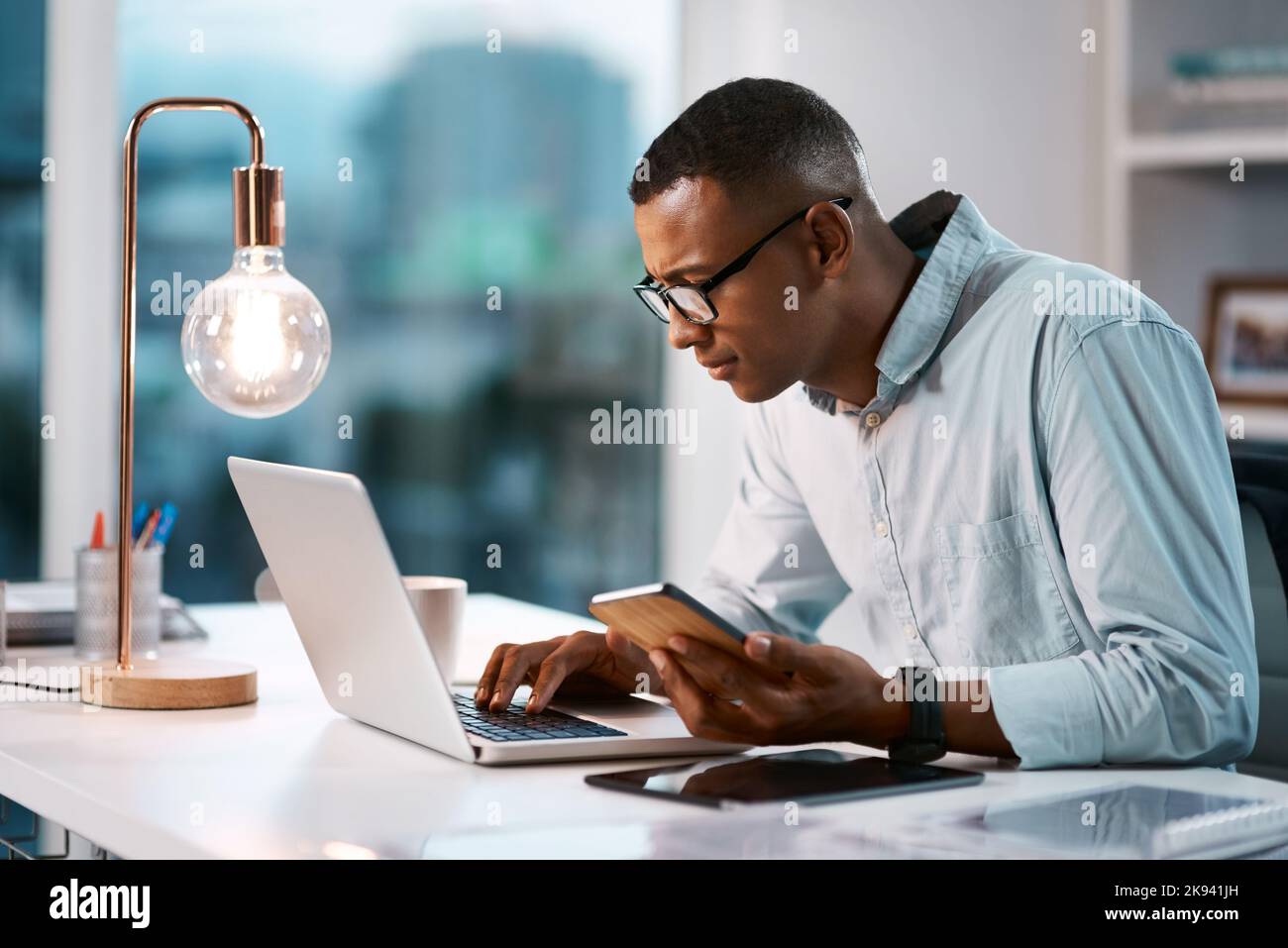 HES è connesso in tutto il mondo degli affari. Un bel giovane uomo d'affari che usa il suo laptop e il cellulare mentre lavora tardi nel suo ufficio. Foto Stock