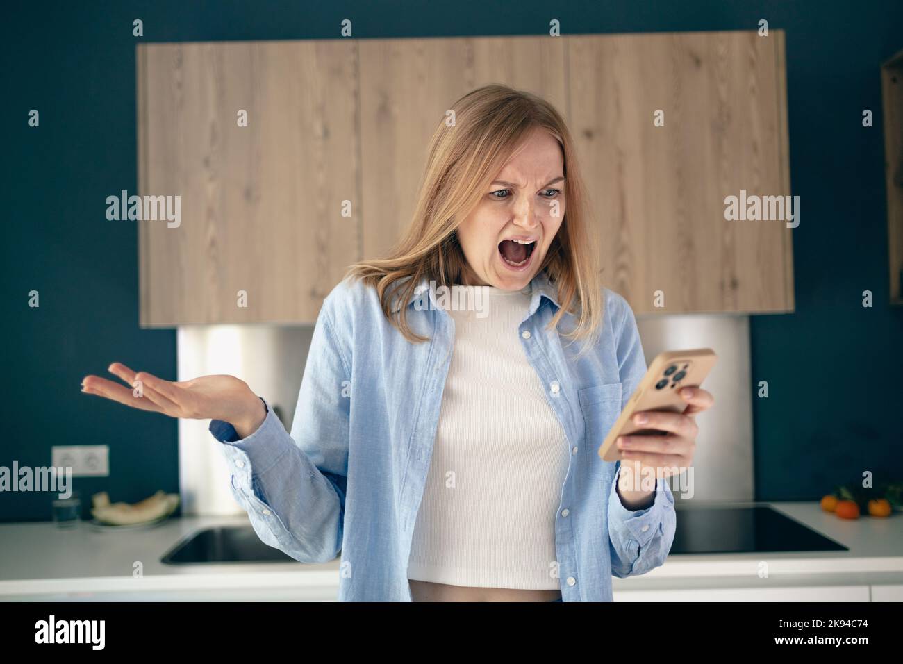 Donna arrabbiata confusa che ha problemi con il telefono in cucina a casa, giovane donna infelice che guarda lo schermo, insoddisfatto per dimesso o rotto Foto Stock