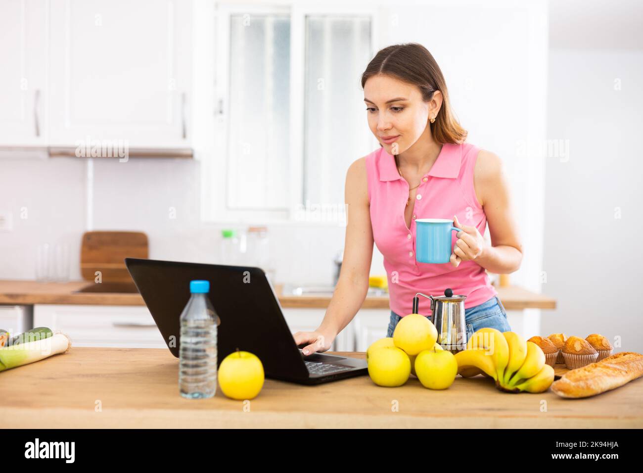 Positiva giovane donna che beve caffè in cucina e che usa il computer portatile Foto Stock