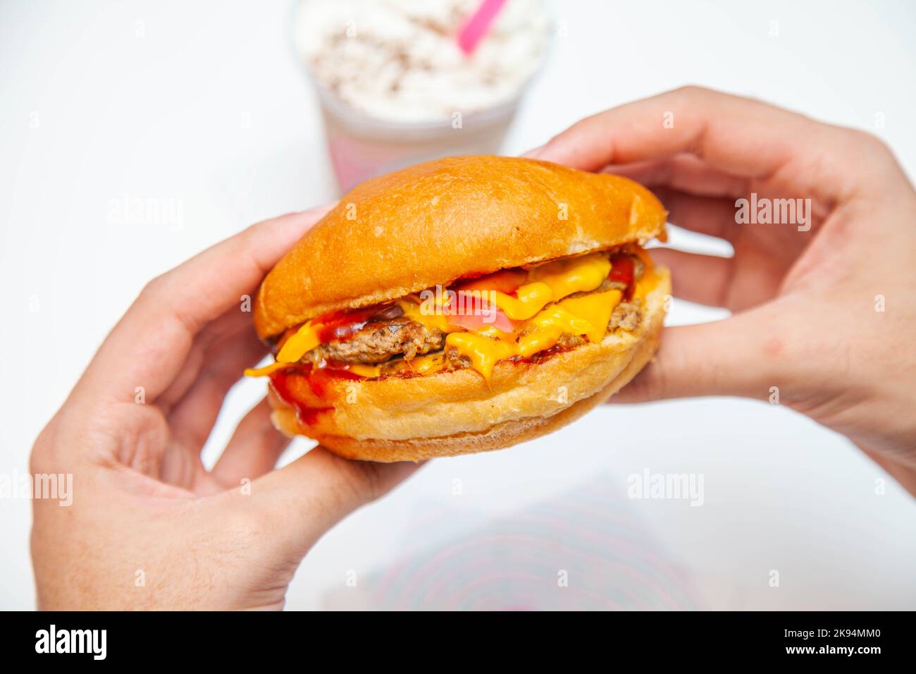 Cheeseburger americano in mano all'interno di un caffè con frappe freddo in sottofondo. Foto Stock