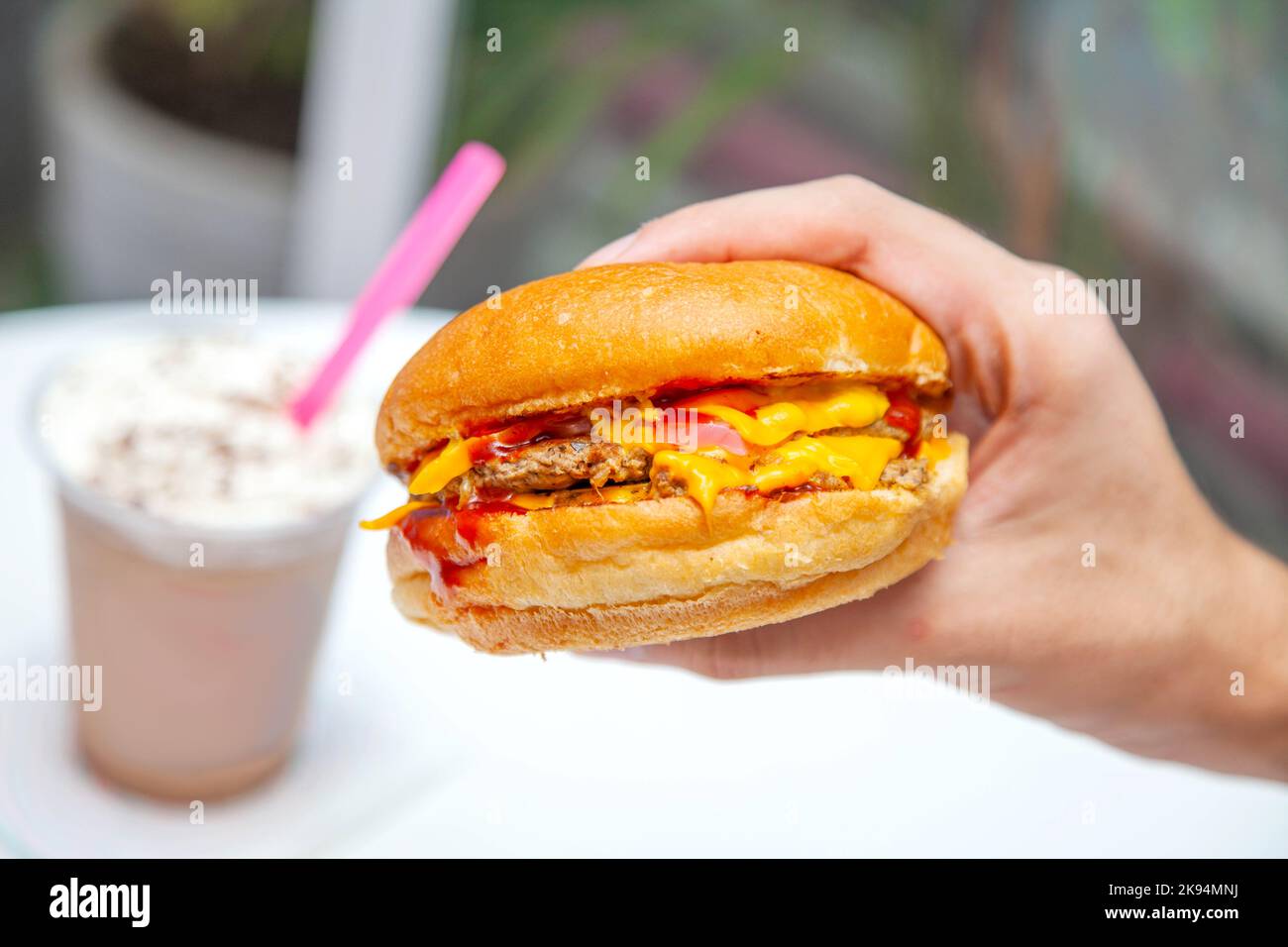 Cheeseburger americano in mano all'interno di un caffè con frappe freddo in sottofondo. Foto Stock