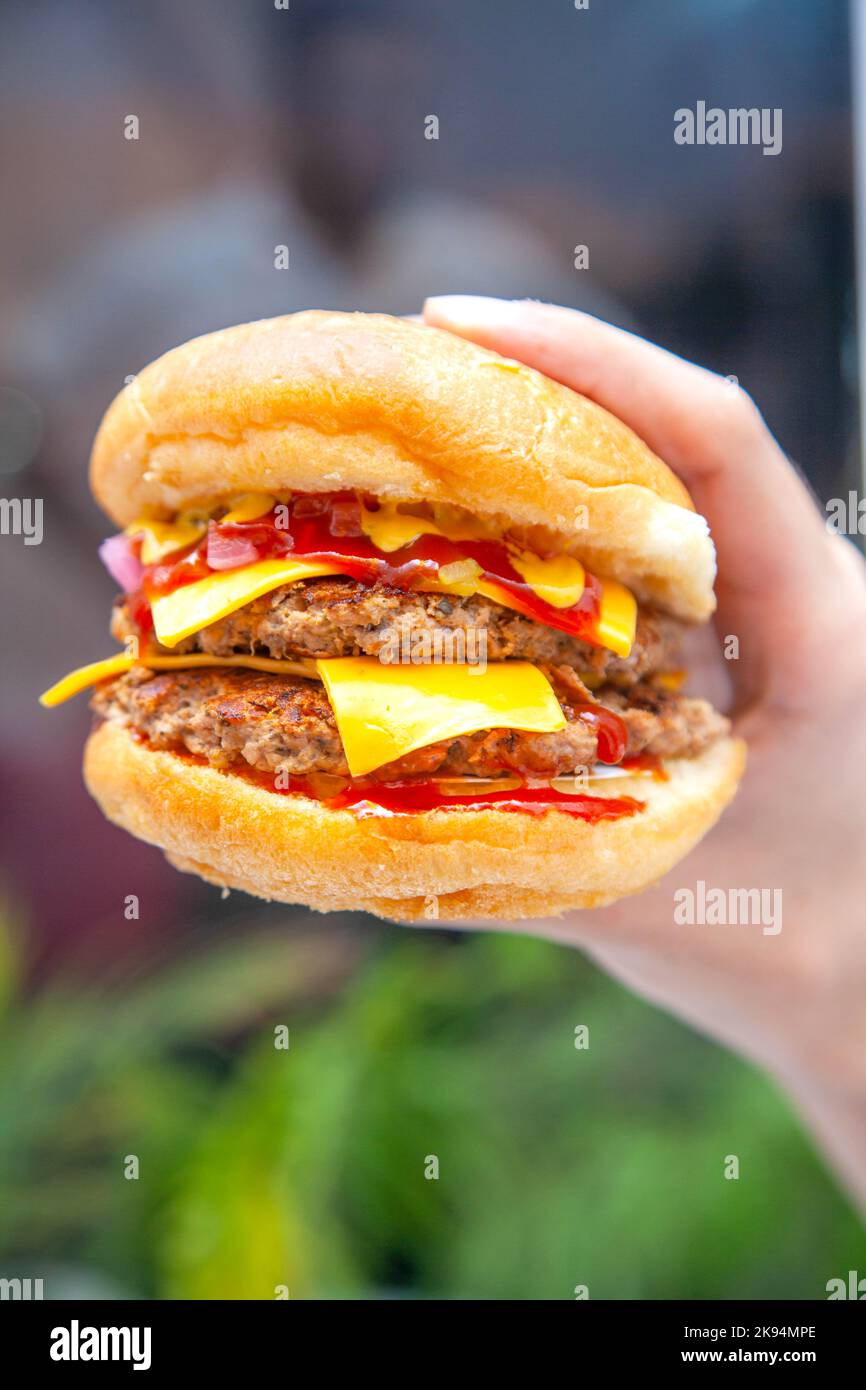 Hamburger di formaggio con tenuta a mano su un lussureggiante sfondo verde. Foto Stock