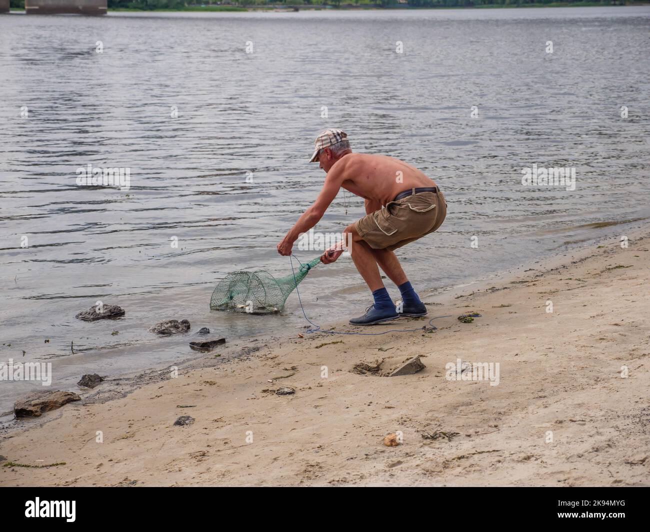 Un vecchio uomo in pantaloncini corti e un cappello e un cavalcetto di pesca in mano che tira il pesce fuori dal fiume Dnipro su una spiaggia urbana vicino al ponte Nord nella città di Kyiv. Foto Stock