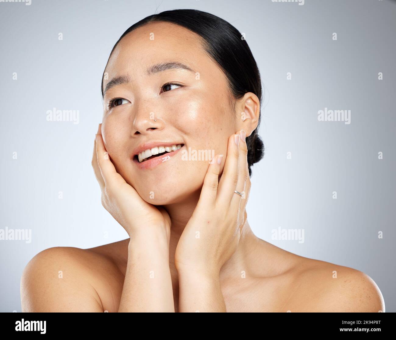 Donna asiatica, viso incandescente e cura della pelle routine con mani che toccano guance su sfondo grigio studio. Sorriso, felice o modello di bellezza in benessere sano Foto Stock